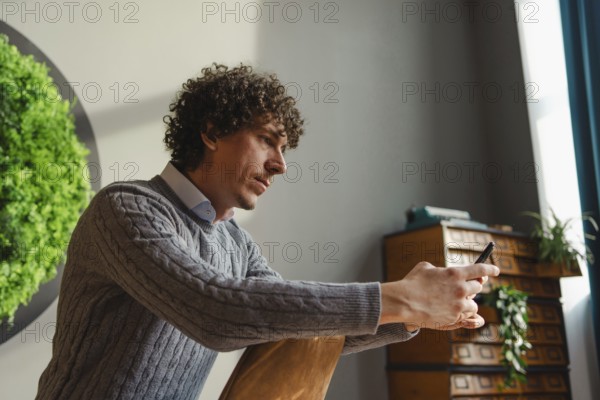 A young man with curly hair uses a smartphone, sitting beside a vibrant green wall. This scene