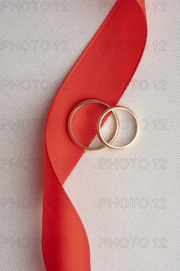 Two elegant gold wedding rings lying on a bold red ribbon, captured on a textured white backdrop. A