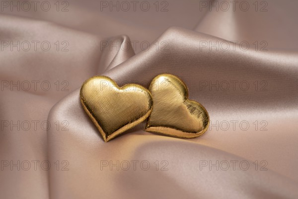 Two golden heart-shaped objects rest gently on a smooth, silky fabric, symbolizing love and