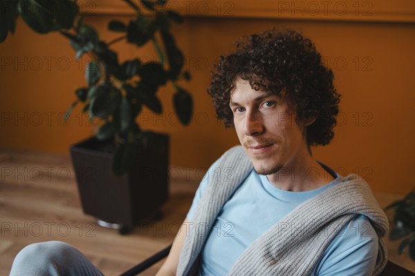 Young man with curly hair in a cozy setting, wearing a blue shirt and a knit sweater. Warm-toned