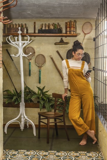 A young woman dressed in fashionable yellow overalls and glasses uses a smartphone while standing