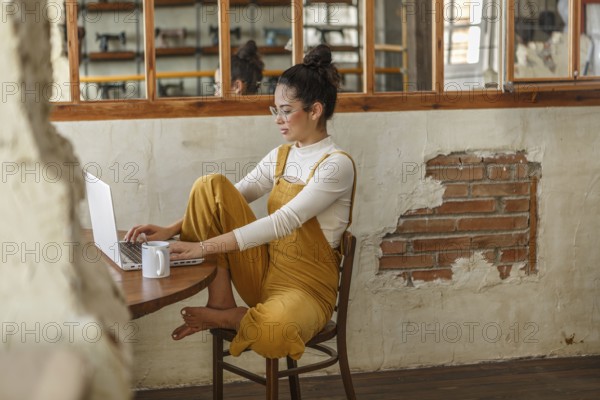 A young woman in a stylishly rustic space focuses on her laptop screen while teleworking, her