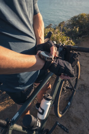 Cropped unrecognizable male cyclist leaning on his bicycle, looking at his watch, with a scenic