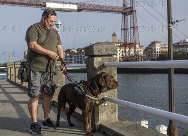 A blind man walks with his guide dog on a riverside path, showcasing companionship and
