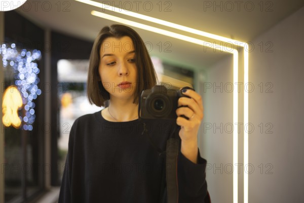 A woman capturing a self-portrait using a DSLR camera in a well-lit interior with neon accents and