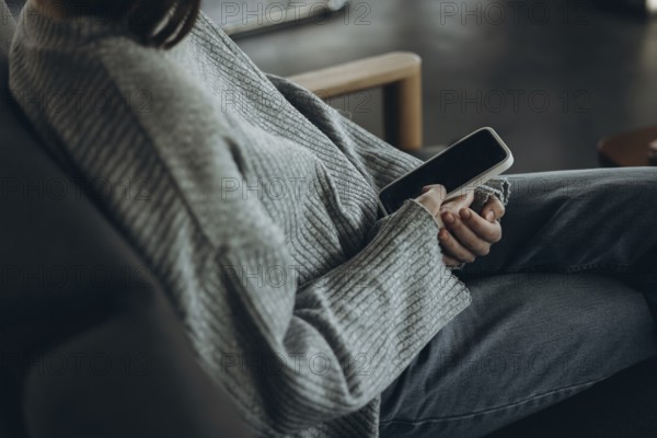 A person wearing a comfy sweater and jeans relaxes on a sofa, holding a smartphone. The setting