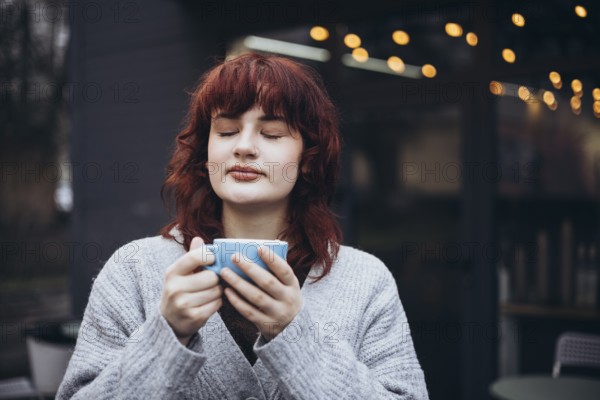 A young woman with red hair, in a gray sweater, enjoys a coffee on cafe terrace. String lights