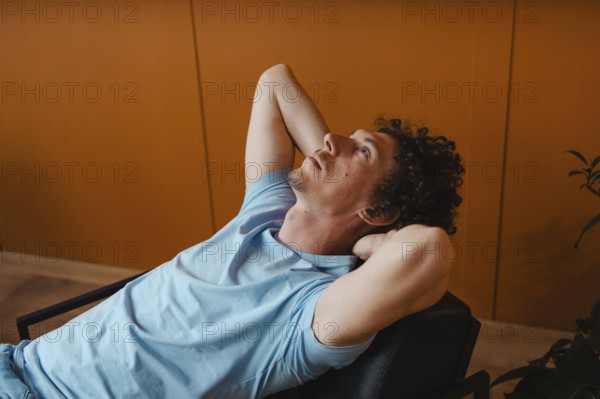 A curly-haired man leans back in a chair with his hands behind his head, gazing upward. The warm
