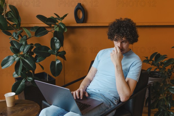 A curly-haired young man in a light blue shirt works on his laptop in a stylish room with plants
