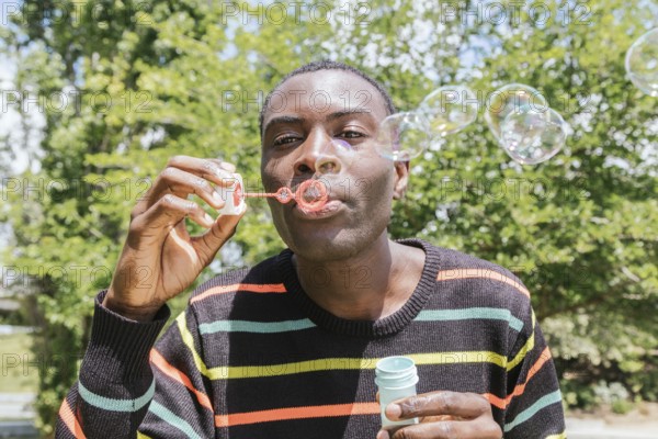 A man in a striped sweater joyfully blows bubbles outside on a bright sunny day, surrounded by lush