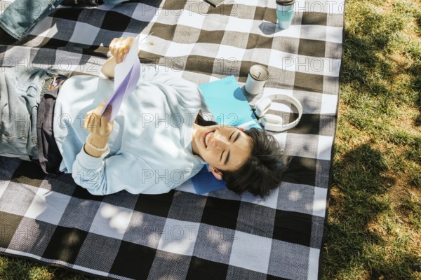 Friends lies on a checkered picnic blanket, enjoying a sunny day with headphones, papers, and