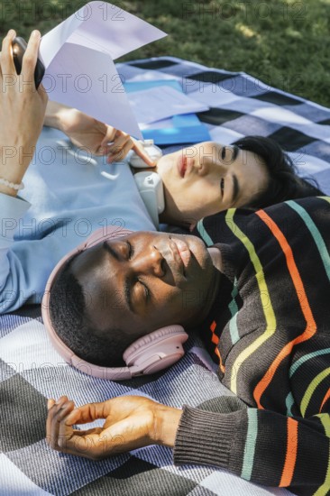 Friends relax outdoors on a sunny day, lying on a blanket. One listens to music with headphones,