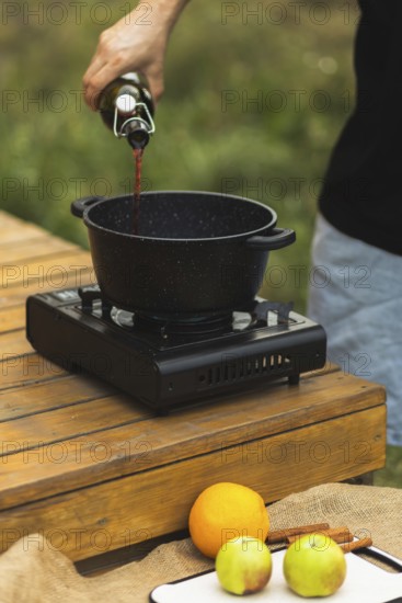 Cropped unrecognizable person pours red wine into a pot for making mulled wine outdoors, surrounded