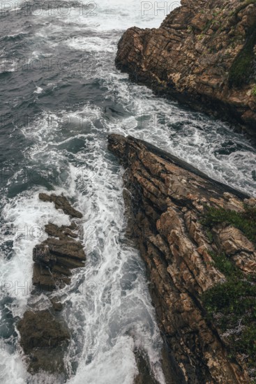A dramatic aerial view of a rocky coastline where powerful ocean waves crash against rugged cliffs.
