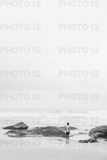 A lone individual stands on a beach surrounded by rocks, gazing at the ocean. The black and white