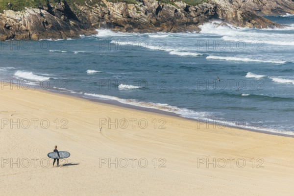A surfer carrying a surfboard walks alone on a sandy beach toward the ocean, with waves crashing