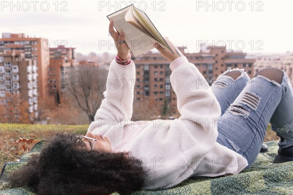 A woman lies on a blanket in a park, reading a book. They wear casual clothes, including jeans and