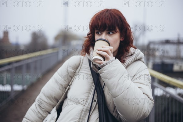A young woman in a warm winter coat drinks coffee from a disposable cup in a day of travel by