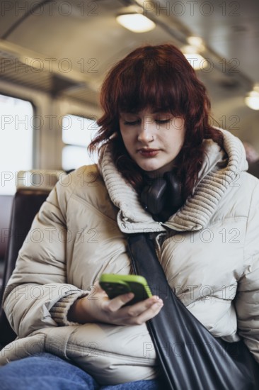 A young woman in a cozy coat, with headphones around her neck, focuses on her smartphone while