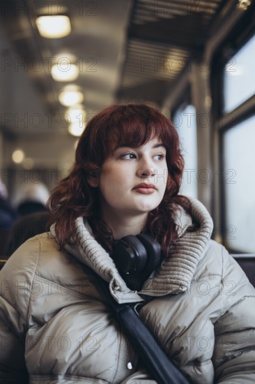 A young woman in a cozy winter coat sits on a train, gazing thoughtfully out the window. Headphones