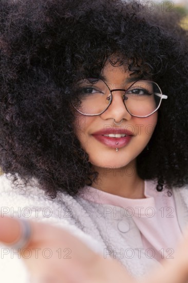 A young latin woman with voluminous curly hair, wearing round glasses and a cozy sweater, smiles