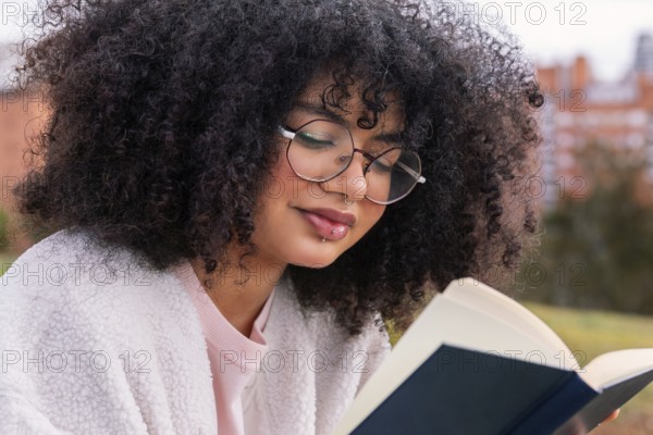 A young latin woman with curly hair and glasses reads a book outdoors, enjoying a peaceful and