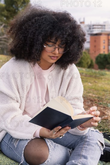 A latin woman with curly hair and glasses enjoys reading a book while seated on grass in a peaceful