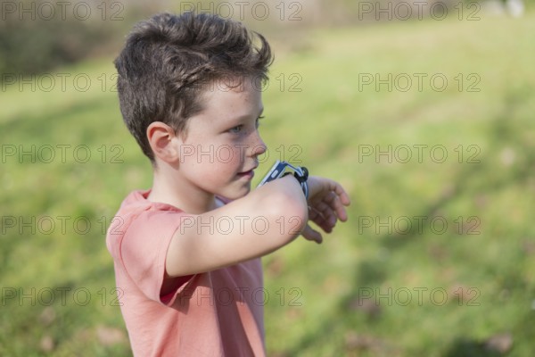 A young boy in a coral shirt examines his smartwatch in a sunny park. The background features