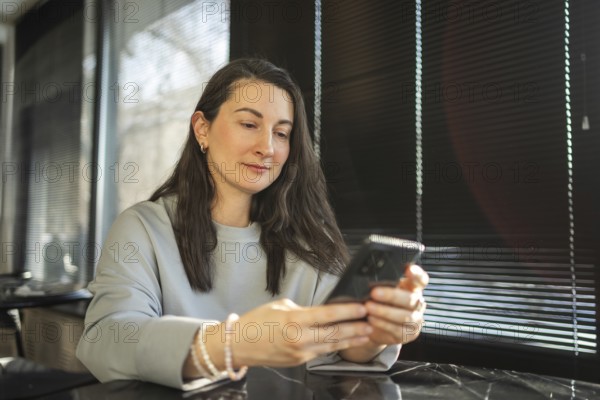 A mid-aged woman is casually using her smartphone in a sunlit cafe environment, reflecting a moment