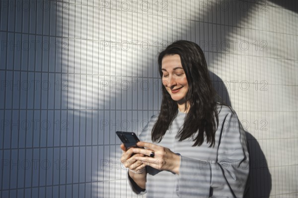 A mid-aged woman enjoys the sunlight as she smiles and engages with her smartphone outdoors against