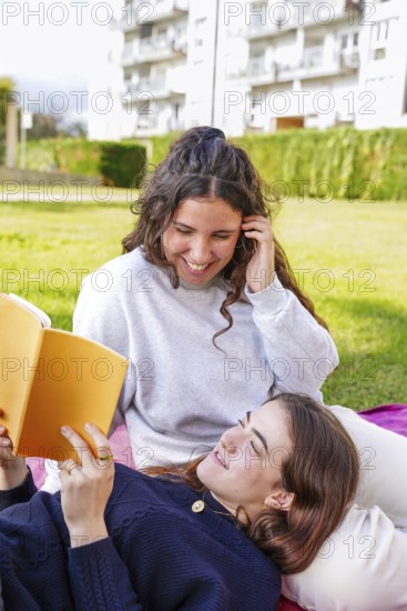 A young women smiles while lying down looking at her partner, who is reading from an orange book in