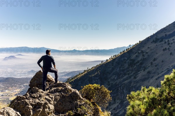 A person stands triumphantly on a rocky mountain peak, gazing over a vast, sunlit valley with