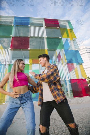 A joyful lesbian couple dances and laughs together in an urban setting. The vibrant glass wall in