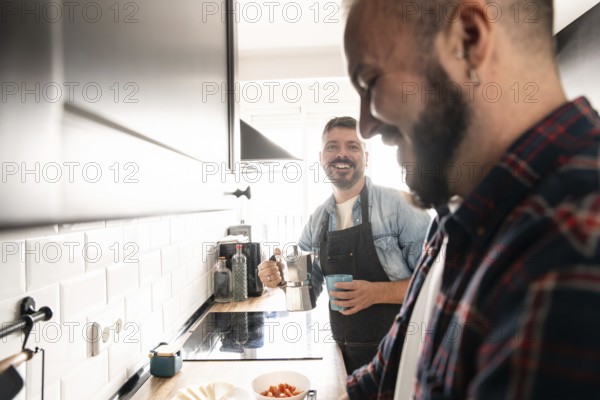 Two gay men enjoy preparing a meal together in a modern kitchen. One holds a coffee pot while the