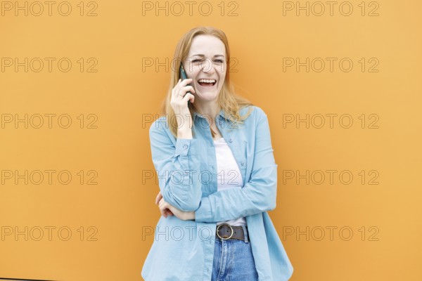 A cheerful woman in a blue shirt talks on her smartphone against an orange background in Warsaw,