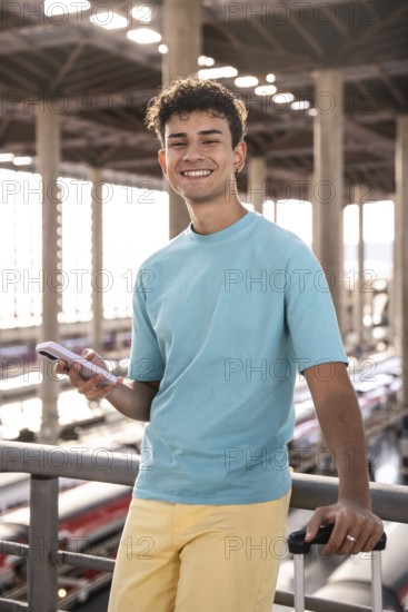 A cheerful young man stands at a train station, holding a phone and a suitcase. He is smiling and