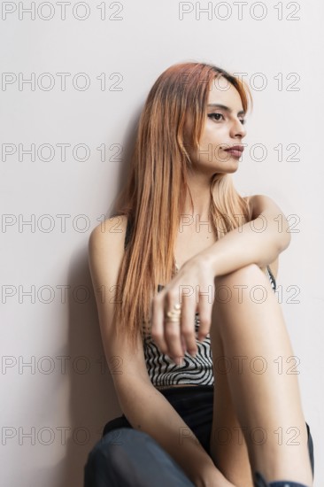 A woman with long hair sits against a white wall, looking contemplative. The close-up captures her