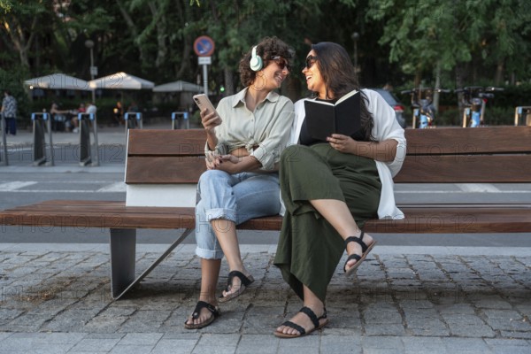 A lesbian couple enjoying a sunny park day. One listens to music with headphones and holds a phone,