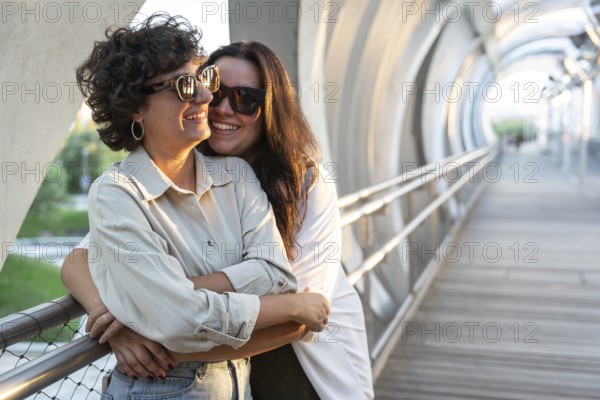 Lesbian couple warmly embracing while standing on a sunlit street bridge. Both wearing sunglasses,