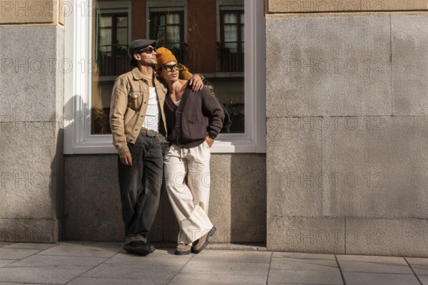 A Colombian gay couple in casual outfits enjoys a sunny urban setting. They lean against a