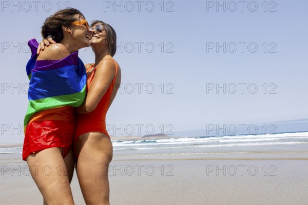 Two lesbians women embrace on a sunny beach, wrapped in a rainbow pride flag, symbolizing love and