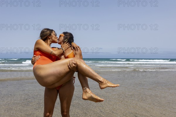 A lesbian couple in swimsuits shares a joyful embrace and kiss on a sunny beach, capturing a moment