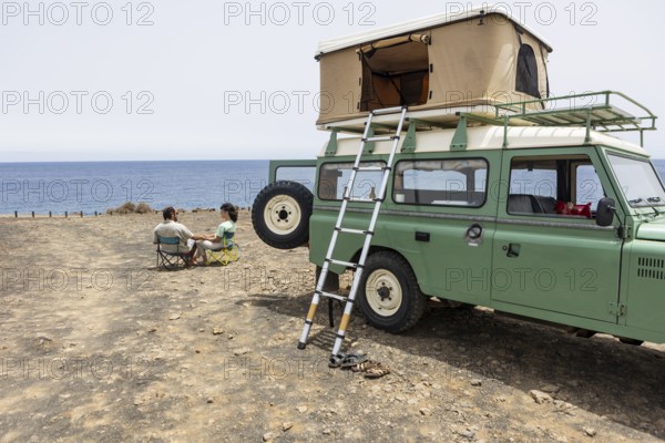 A blind man and a woman sit on camping chairs near their motorhome, overlooking the ocean. The