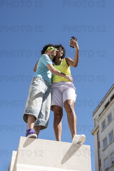 A joyful, multiethnic LGBTQ+ couple captures a selfie outdoors. They stand closely on a sunny day,