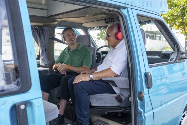 A young man and an elderly man in a wheelchair share a joyful moment inside a blue camper van. The