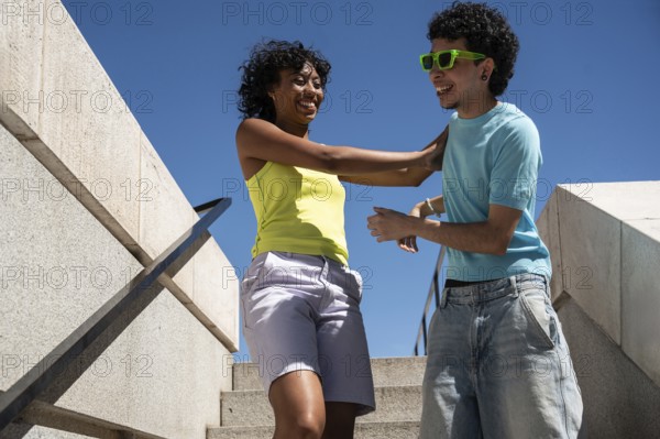 A vibrant, multiethnic LGBTQ+ couple shares a joyful moment on outdoor steps. Both dressed in