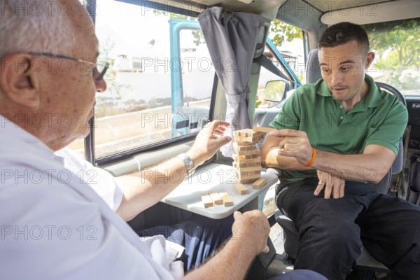 An elderly man and his adult son enjoy playing a game of Jenga inside a van, parked in a sunny