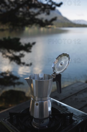 A moka pot sits on a camping stove beside a tranquil Norwegian lake, surrounded by lush mountains