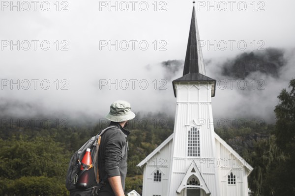 A traveler with a backpack and hat stops in front of a picturesque white Norwegian church,