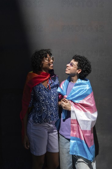 A joyful multiethnic LGBTQ+ couple smiling at each other, wrapped in Pride flags. Captured in